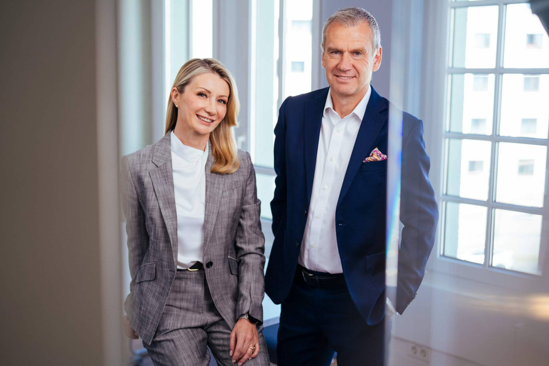 Foto: Zwei lächelnde Geschäftspartner – ein Mann im blauen Anzug mit verschränkten Armen und eine Frau in weißer Bluse und grauer Hose – stehen nebeneinander in einem hellen Büro