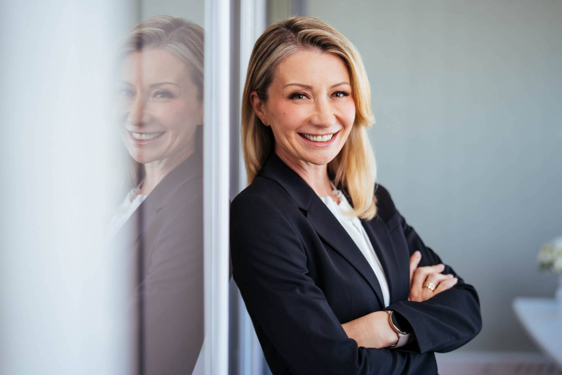 Foto: Lächelnde blonde Geschäftsfrau im schwarzen Blazer, mit verschränkten Armen an eine Glaswand gelehnt. Portrait von Carolin Bucherer