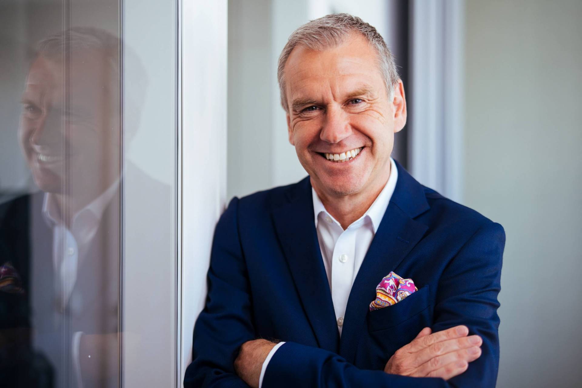 Foto: Lächelnder Geschäftsmann im blauen Anzug mit buntem Einstecktuch, mit verschränkten Armen an eine Glaswand gelehnt.