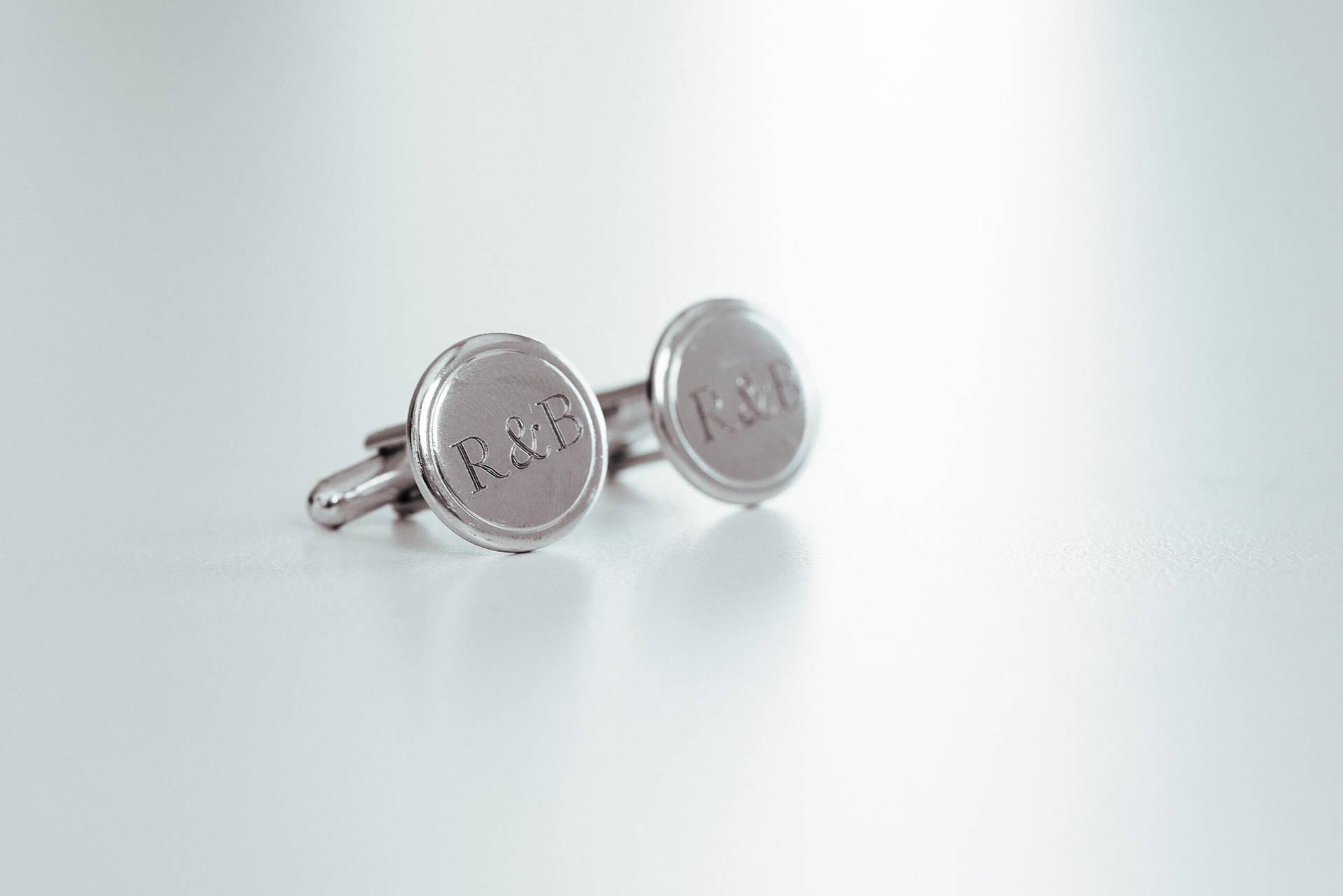 Foto: Zwei silberne Manschettenknöpfe mit eingravierten Buchstaben ‚R & B‘ auf hellem Untergrund.