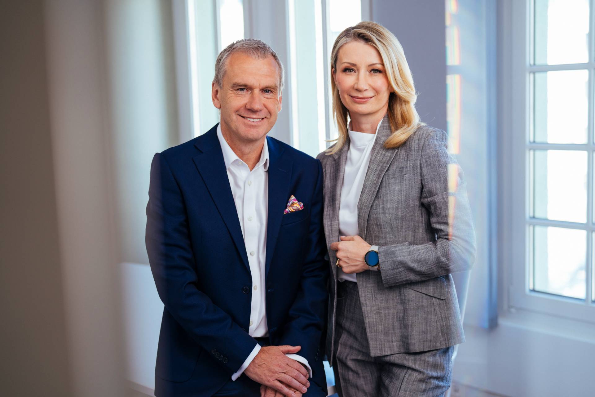 Foto: Zwei lächelnde Geschäftspartner – eine Frau im grauen Hosenanzug mit weißer Bluse und ein Mann im blauen Anzug mit Einstecktuch – stehen nebeneinander vor einem großen Fenster in einem hellen Büro.